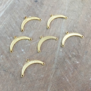 May include: Six gold-colored metal crescent moon shapes with three holes each. The moon shapes are arranged in a semi-circle on a wooden surface.