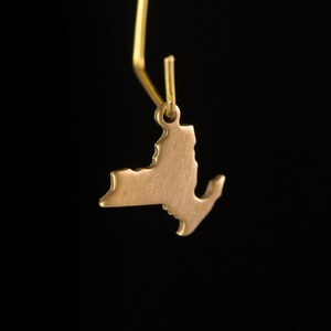 May include: A gold-colored pendant shaped like the state of New York. The pendant is hanging from a gold-colored chain.