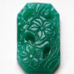 Puede incluir: Un botón de vidrio verde con un diseño floral tallado. El botón tiene una forma ligeramente irregular y mide aproximadamente 2,5 cm de diámetro.
