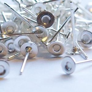 May include: A close-up shot of a pile of silver-colored earring posts with flat, circular tops. The posts are scattered, with some in focus and others blurred, against a white background. The posts are small, with a simple, minimalist design.