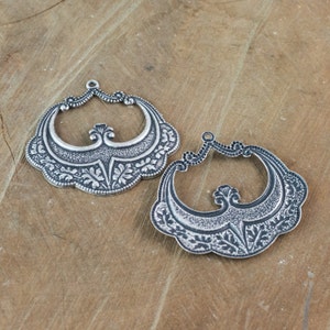 Könnte beinhalten: Zwei silberfarbene Metall-Anhänger mit einem verzierten Design. Jeder Anhänger hat die Form eines Halbmondes mit filigranen Blumendetails und einem zentralen Dekorelement.