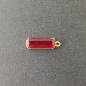 May include: A rectangular, ruby-red gemstone pendant with a gold-coloured frame and a small loop for hanging. The pendant is set against a dark grey background. The gemstone has a faceted appearance.