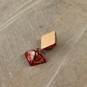 May include: Two red glass dice with gold accents and a floral pattern. The dice are on a wooden surface.