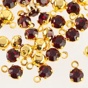 Pode incluir: Close-up de amuletos de metal dourado espalhados com pedras preciosas facetadas de cor vermelha profunda. Cada amuleto tem um pequeno laço para fixação. As pedras são redondas e engastadas em uma moldura dourada.