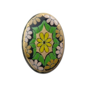 Puede incluir: Botón de forma ovalada con fondo negro y un diseño floral colorido. El centro del botón presenta una flor amarilla rodeada de hojas verdes. El diseño está rodeado de flores blancas y amarillas más pequeñas.