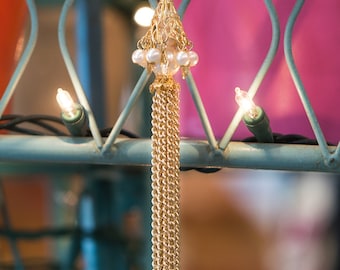 Könnte beinhalten: Ein goldener Quastenanhänger mit einem Design aus weißen Perlen und goldenen Perlen. Der Anhänger hängt an einer Metallkette und ist an einem blauen Metallzaun befestigt.