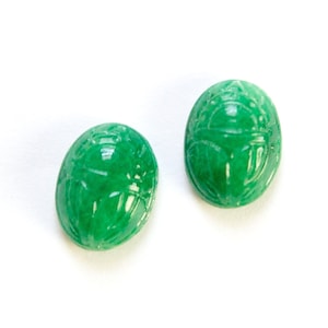 May include: Two bright green, oval-shaped scarab beetle carvings. The carvings have detailed etched patterns and are set against a white background. The scarabs are likely made of jade or a similar stone.