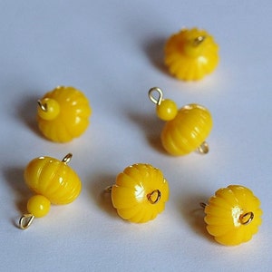 May include: Six bright yellow, pumpkin-shaped beads with gold-colored loops for jewelry making. The beads have a ribbed texture and a small, round bead attached to some of them.