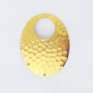 Puede incluir: Un colgante de metal ovalado de color dorado con textura martillada y un recorte circular en el centro. El colgante tiene cuatro agujeros alrededor del borde para sujetar una cadena o cordón.