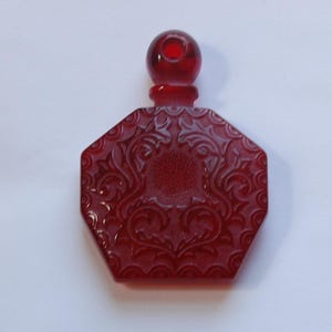 Peut inclure: Un charme de bouteille de parfum en verre rouge avec une base octogonale et un motif floral détaillé. La bouteille a un bouchon rond.