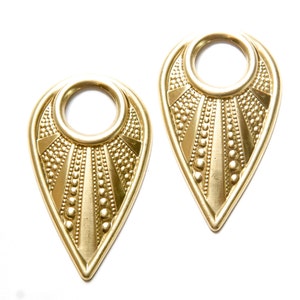 May include: A pair of gold-tone metal earrings with a teardrop shape and a circular cutout in the center. The earrings have a textured, geometric design.
