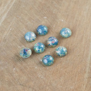 Könnte beinhalten: Acht runde, blau-grüne Cabochons mit gesprenkeltem, schillerndem Aussehen. Die Cabochons haben eine flache Rückseite und eine gewölbte Oberseite, verstreut auf einer hellbraunen Holzoberfläche.
