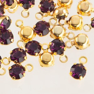 May include: Close-up of gold-colored metal jewelry findings with deep purple, round gemstones. Each gem is set in a gold bezel and connected to a small loop for attaching to other components. The findings are scattered across a white surface.