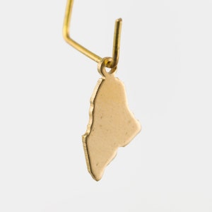 May include: A gold-colored pendant shaped like the outline of a country. The pendant is attached to a gold-colored jump ring.