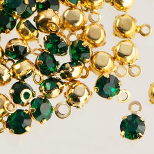 May include: Close-up of emerald green gemstone beads with gold-colored metal settings. The beads have a round, faceted cut and are scattered across a white surface. Each bead has a small loop for stringing or attaching.