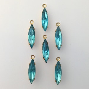 May include: Six aquamarine-colored, marquise-shaped crystal pendants with gold-tone metal settings and loops. The crystals are a vibrant teal blue, and the metal has a warm, antique finish. These pendants are ideal for jewelry making.