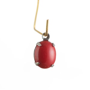 May include: A single red oval gemstone pendant with a silver-colored metal setting and a gold-colored wire. The gemstone is a vibrant red color and is the focal point of the pendant. The setting has four prongs holding the stone.