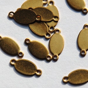May include: Close-up of several small, oval-shaped brass tags with a loop at each end. The tags are scattered across a light-colored surface, with some overlapping. The brass has a slightly aged, vintage appearance.