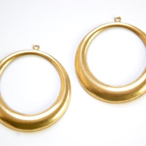 May include: Two gold-colored metal hoops with a smooth, rounded finish. The hoops have a small loop at the top for attaching to a chain or other jewelry.