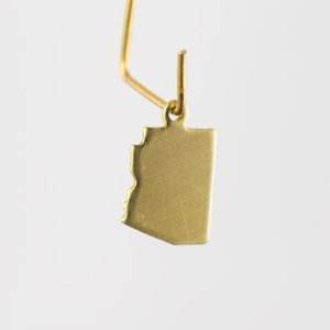 May include: A gold-colored pendant in the shape of the state of Arizona, with a jagged edge representing the state's border. The pendant is suspended from a thin, gold-colored wire, creating a minimalist and elegant design.