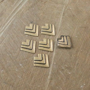 May include: Six gold-coloured metal heart-shaped charms with a geometric pattern. The charms are arranged in a triangular shape on a wooden surface.