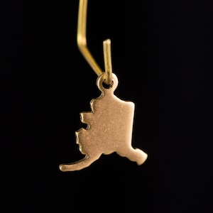 May include: A gold charm in the shape of the state of Alaska. The charm is attached to a gold chain.