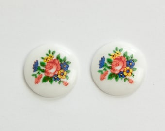 May include: Two white, round cabochons with a floral design. Each cabochon features a central red rose surrounded by blue, yellow, and green floral accents. The cabochons are likely for jewelry making or craft projects.