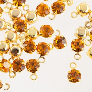 May include: Close-up of scattered golden-colored jewelry findings. Each finding features a round, amber-colored gemstone set in a gold-tone metal setting. The findings have small loops for attachment, ideal for jewelry making and crafting projects.