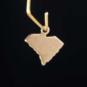 May include: A gold-colored charm in the shape of the state of South Carolina hangs from a gold-colored hook. The charm is suspended against a black background.