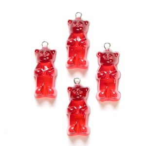 Pode incluir: Quatro pingentes de urso de goma vermelho com laços prateados. Os ursos de goma estão de pé com os braços cruzados.