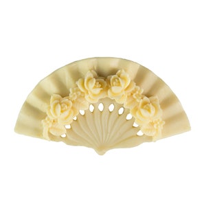 Puede incluir: Un broche blanco en forma de abanico con un diseño tallado de flores. El broche está hecho de un material liso, similar al marfil.