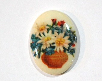 May include: A white oval ceramic tile with a floral design. The tile features a bouquet of white flowers with yellow centers in a brown pot. The flowers are surrounded by green leaves and blue flowers.