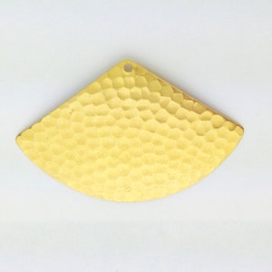 Puede incluir: Un colgante de metal de color dorado con forma de abanico y textura martillada. El colgante tiene un agujero en la parte superior para sujetar una cadena o cordón.