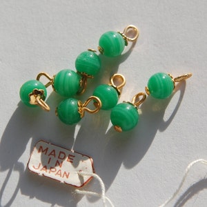 May include: A group of seven green glass beads with gold accents and a small tag that reads "Made in Japan".