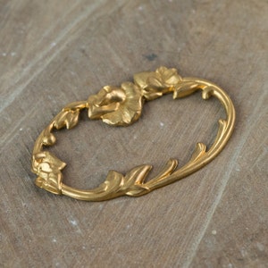 May include: A gold-colored metal decorative trim with a floral design. The trim is shaped like an oval with a leafy design and two flower shapes.