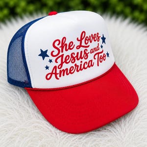 May include: A red, white, and blue trucker hat with the text "She Loves Jesus and America Too." The hat features a red brim, a white front panel with the text, a blue mesh back, and a red button on top.