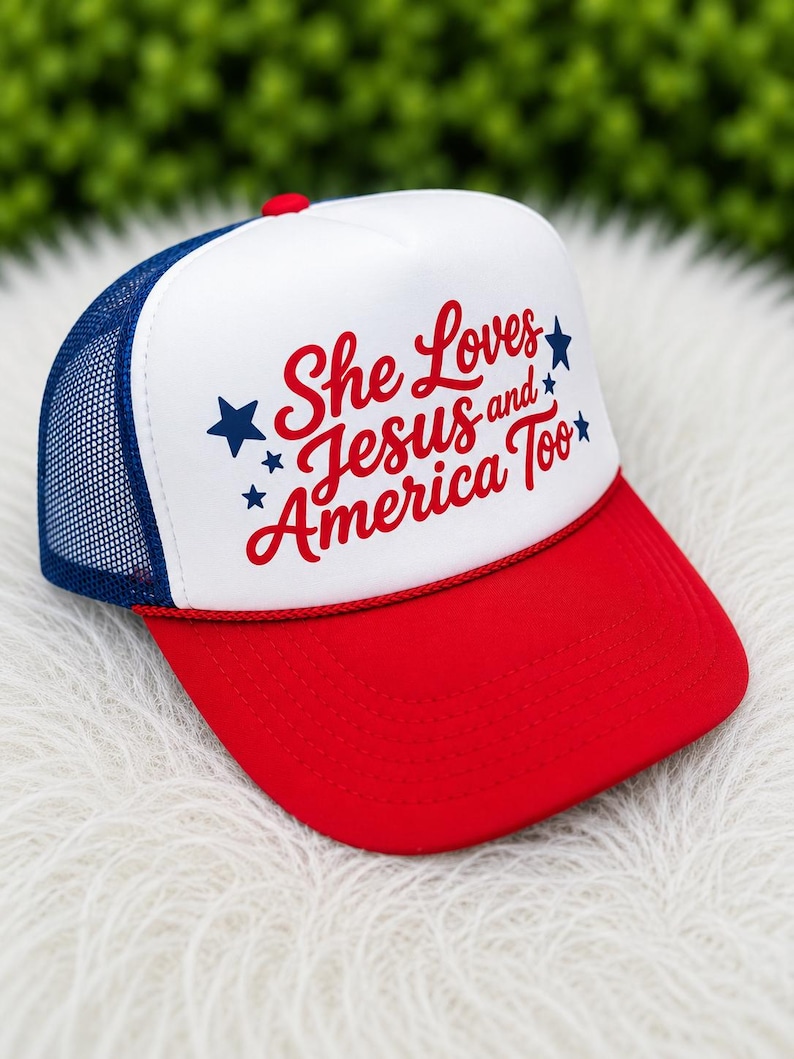 May include: A red, white, and blue trucker hat with the text "She Loves Jesus and America Too." The hat features a red brim, a white front panel with the text, a blue mesh back, and a red button on top.