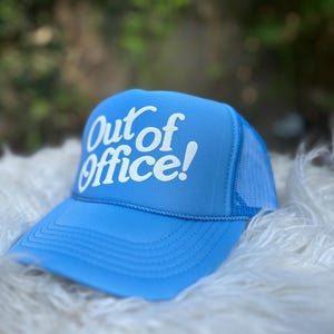 Może przedstawiać: Jasnoniebieska czapka trucker z siateczką z tyłu. Na czapce z przodu widnieje napis "Out of Office!" w kolorze białym.