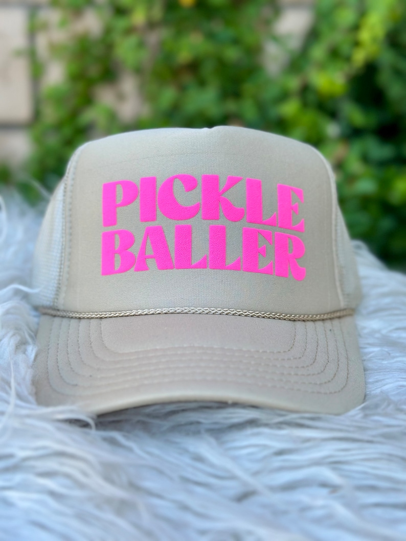 May include: A beige trucker hat with a pink "PICKLE BALLER" logo.