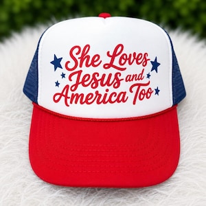 May include: A red, white, and blue trucker hat with the text "She Loves Jesus and America Too." The hat features a red brim, a white front panel with the text, a blue mesh back, and a red top button.
