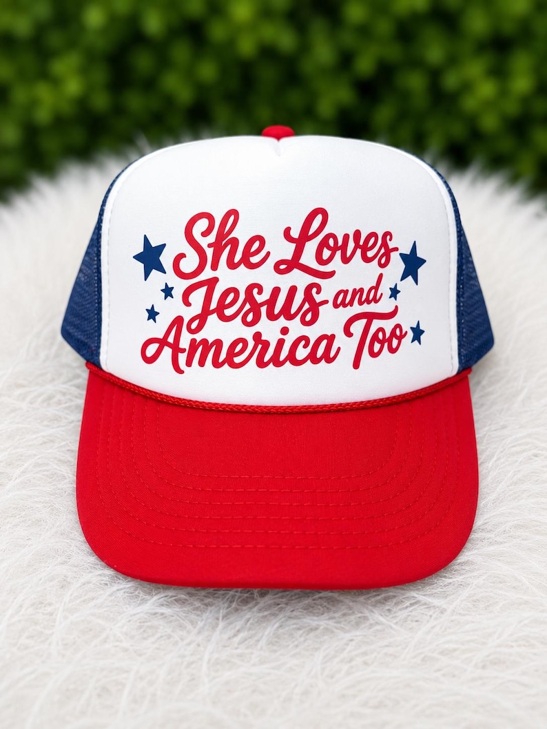 May include: A red, white, and blue trucker hat with the text "She Loves Jesus and America Too." The hat features a red brim, a white front panel with the text, a blue mesh back, and a red top button.