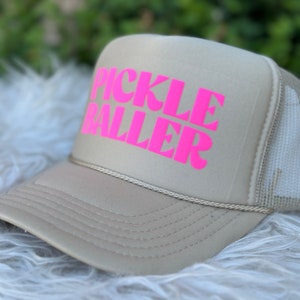 May include: A beige trucker hat with a pink "PICKLE BALLER" logo.