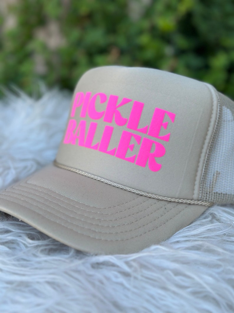 May include: A beige trucker hat with a pink "PICKLE BALLER" logo.