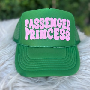 May include: A green trucker hat with the text "PASSENGER PRINCESS" in pink, retro-style lettering. The hat features a mesh back and a curved brim. The hat is sitting on a white, fluffy surface.