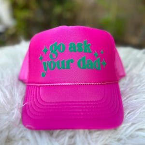 May include: A pink trucker hat with a green embroidered design that says "go ask your dad" with two small stars on either side of the text.