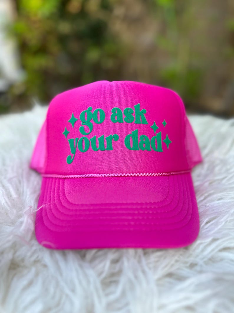K&ouml;nnte beinhalten: Eine rosafarbene Trucker-Kappe mit einem gr&uuml;nen gestickten Design, auf dem "go ask your dad" steht, mit zwei kleinen Sternen auf jeder Seite des Textes.