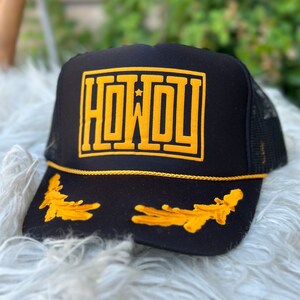 May include: Black trucker hat with a gold embroidered "HOWDY" and a gold star on the front. The hat also has gold embroidered leaves on the brim.