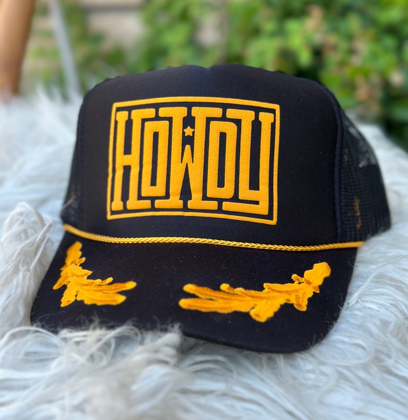 May include: Black trucker hat with a gold embroidered "HOWDY" and a gold star on the front. The hat also has gold embroidered leaves on the brim.