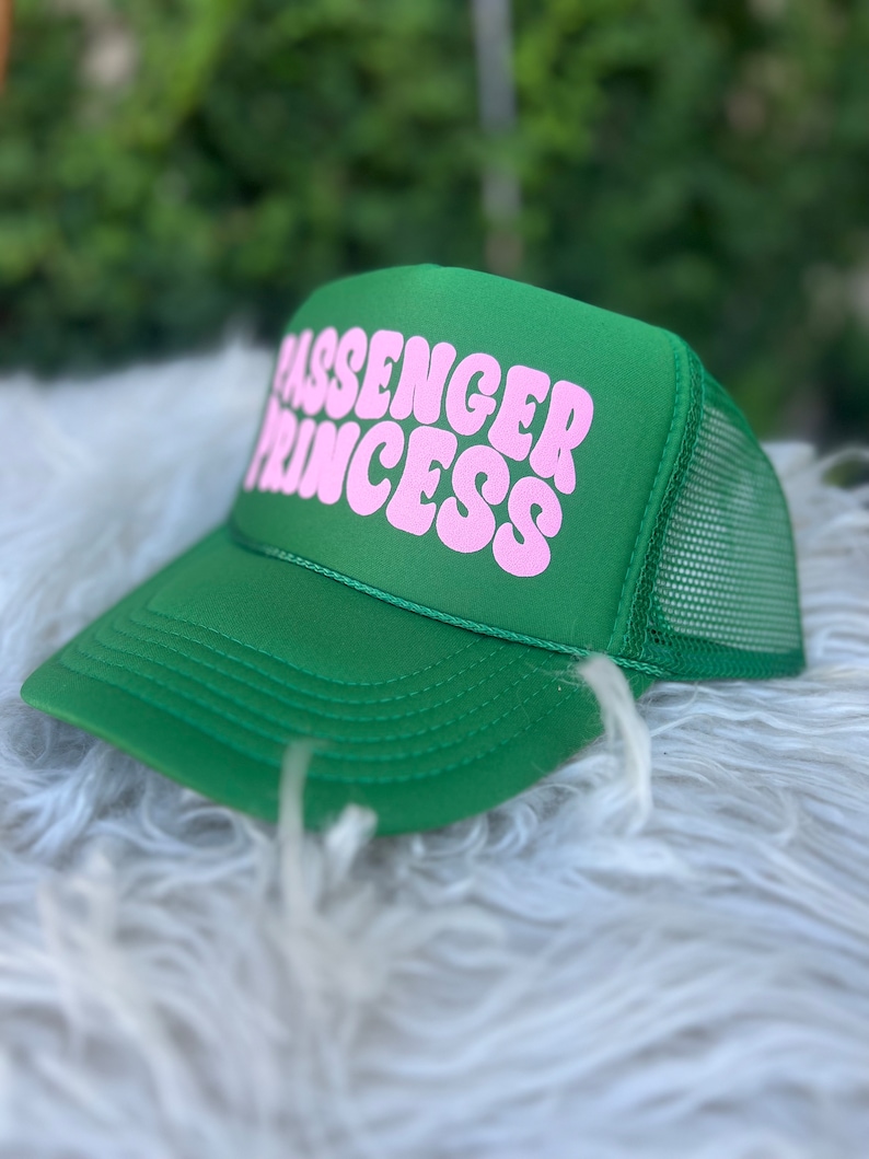 May include: A green trucker hat with the words "PASSENGER PRINCESS" in pink, block letters. The hat has a mesh back and a curved brim. The hat is sitting on a white, fluffy surface.
