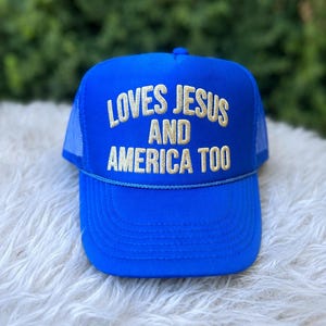 May include: A blue trucker hat with the embroidered text "LOVES JESUS AND AMERICA TOO" in gold thread. The hat has a mesh back and a curved brim. The hat is sitting on a white, fluffy surface.